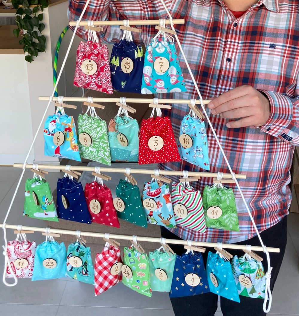 Eine Person im karierten Hemd hält einen selbstgemachten Adventskalender hoch. Der Adventskalender besteht aus Holzstäben und weißer Kordel und hat die Form eines nach oben spitz zulaufenden Tannenbaums. An den Holzstäben sind Stoffsäckchen in den Farben Grün, Rot und Blau mit Holzklammern befestigt. An jedem Stoffsäckchen hängt ein runder Holzanhänger mit einer Zahl.