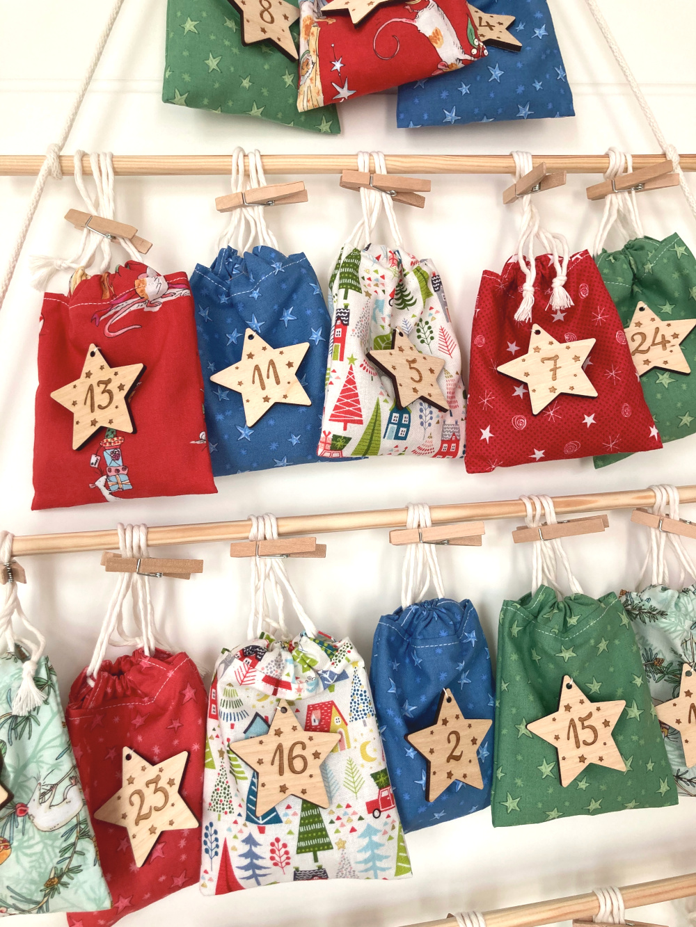 Selbstgemachter Adventskalender: Zu sehen sind rote, blaue, grüne und bunt bedruckte Stoffsäckchen, die mit Holzklammern an Stäben befestigt sind. An den Säckchen hängen Holzanhänger in Form eines Sterns. Auf den Holzanhängern stehen Zahlen von 1 bis 24.