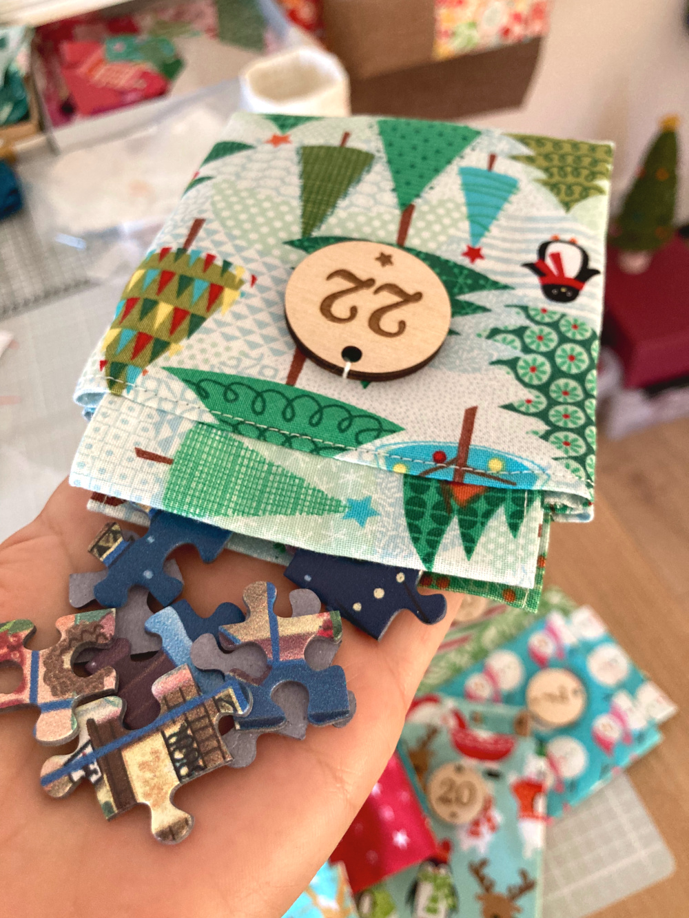 Eine flache Hand, auf der ein Adventskalendersäckchen und ein paar Puzzleteile liegen. Auf dem Säckchen sind Weihnachtsbäume in verschiedenen Grüntönen abgebildet, und es hängt ein runder Holzanhänger mit der Zahl 22 daran.