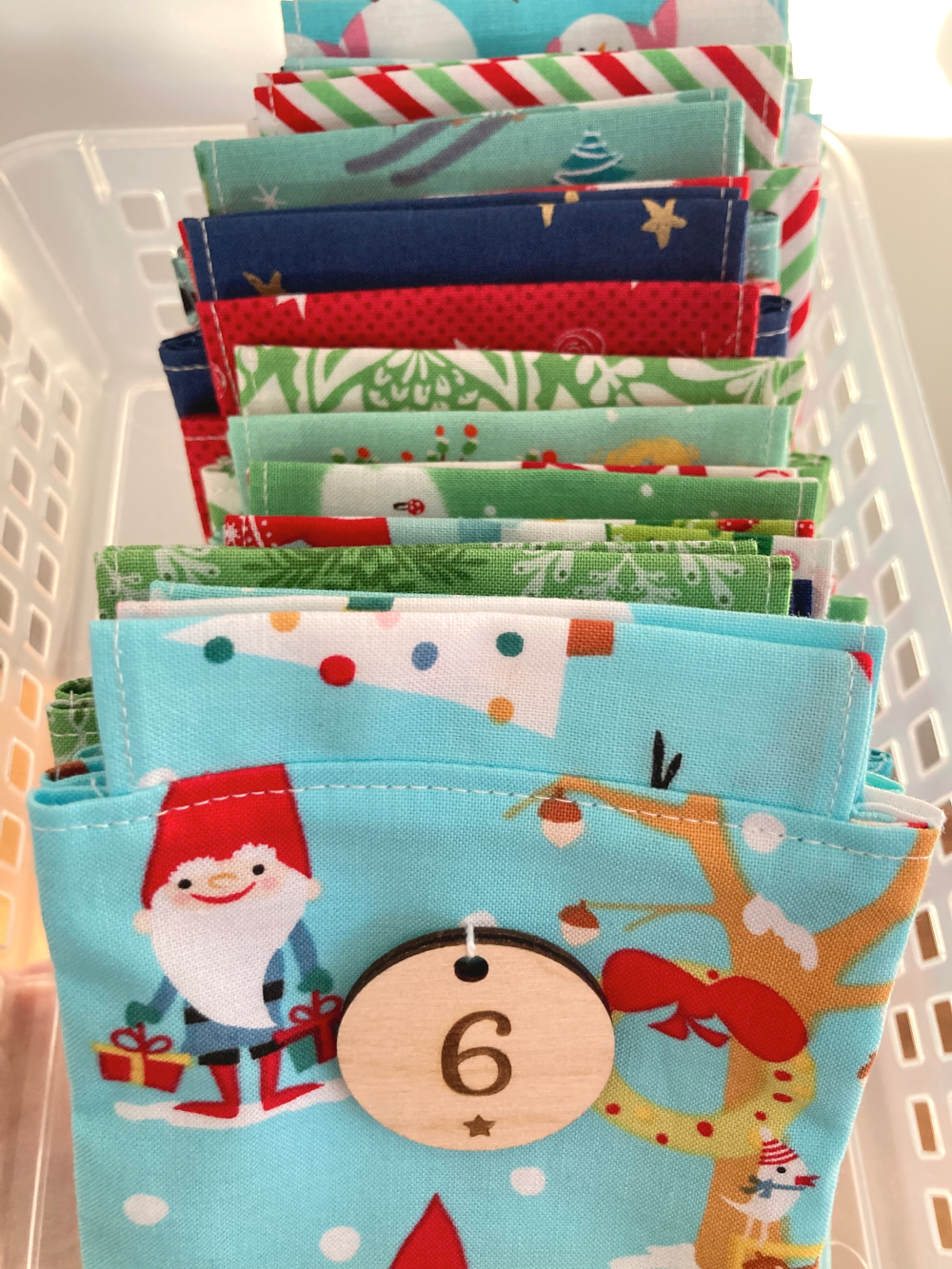 Adventskalendersäckchen aus grünen, roten und türkisblauen Weihnachtsstoffen, hintereinander aufgestellt. Ganz vorne ein türkisfarbenes Säckchen, auf dem zwei Weihnachtswichtel mit roter Mütze und weißem Bart abgebildet sind. Am Säckchen hängt ein runder Holzanhänger mit der Zahl 6.