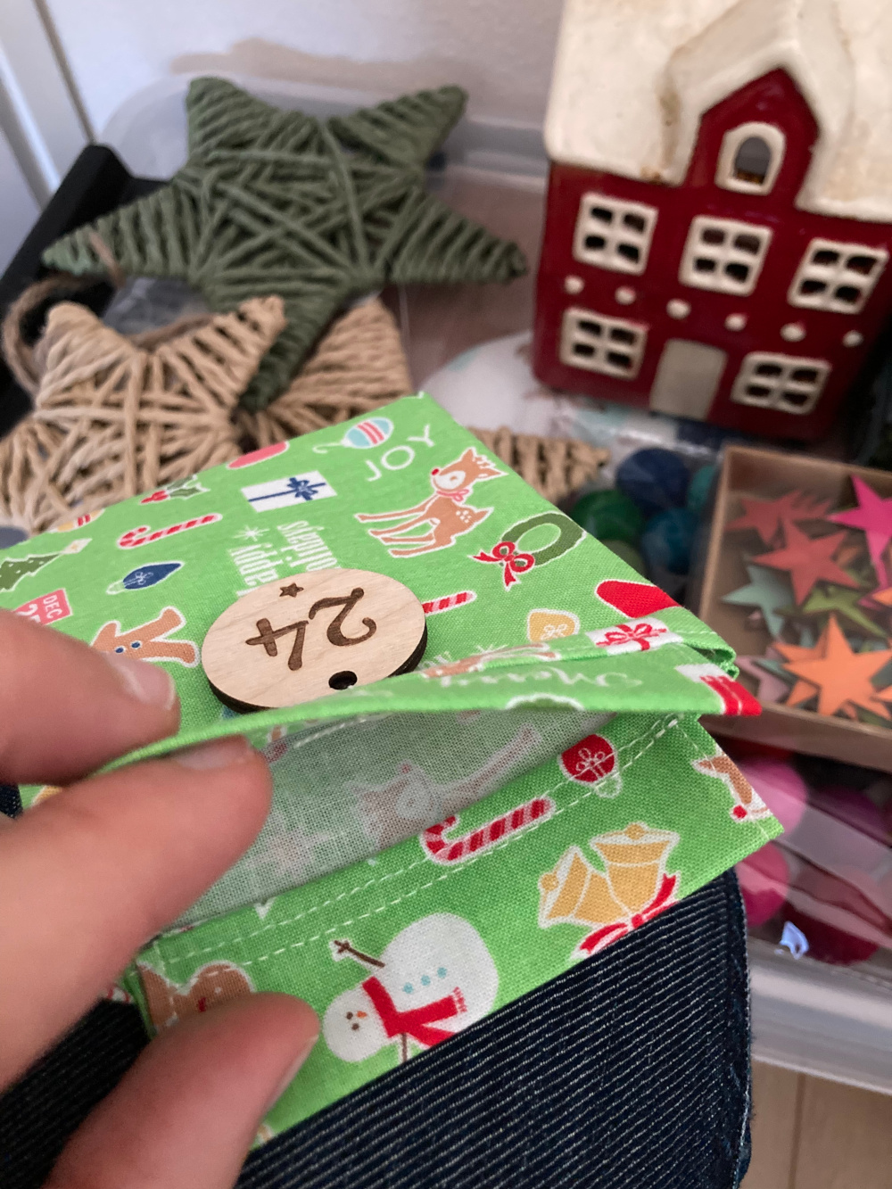 Adventskalendersäckchen aus grünem Weihnachtsstoff mit eine rundem Holzanhänger, in den die Zahl 24 eingraviert ist. Zeigefinger und Daumen einer Hand öffnen das Säckchen, damit man die Naht im Inneren sehen kann. Im Hintergrund verschiedene Weihnachtsdeko: Sterne und ein rotes Keramikhaus.