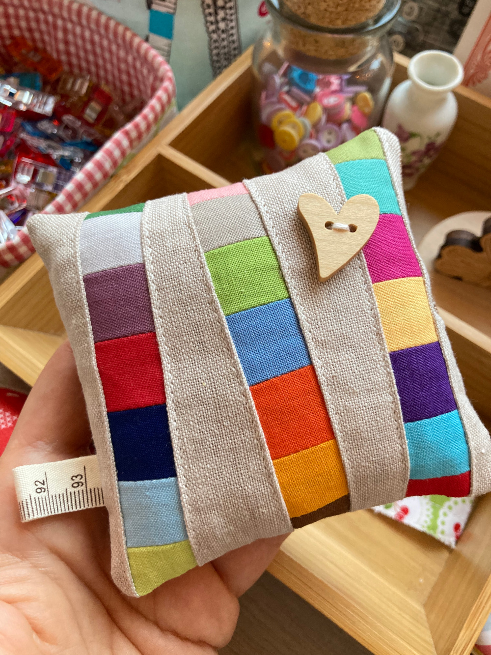 Improv Patchwork: Nadelkissen aus grauem Leinenstoff mit bunten Quadraten und aufgenähtem Herz-Holzknopf.