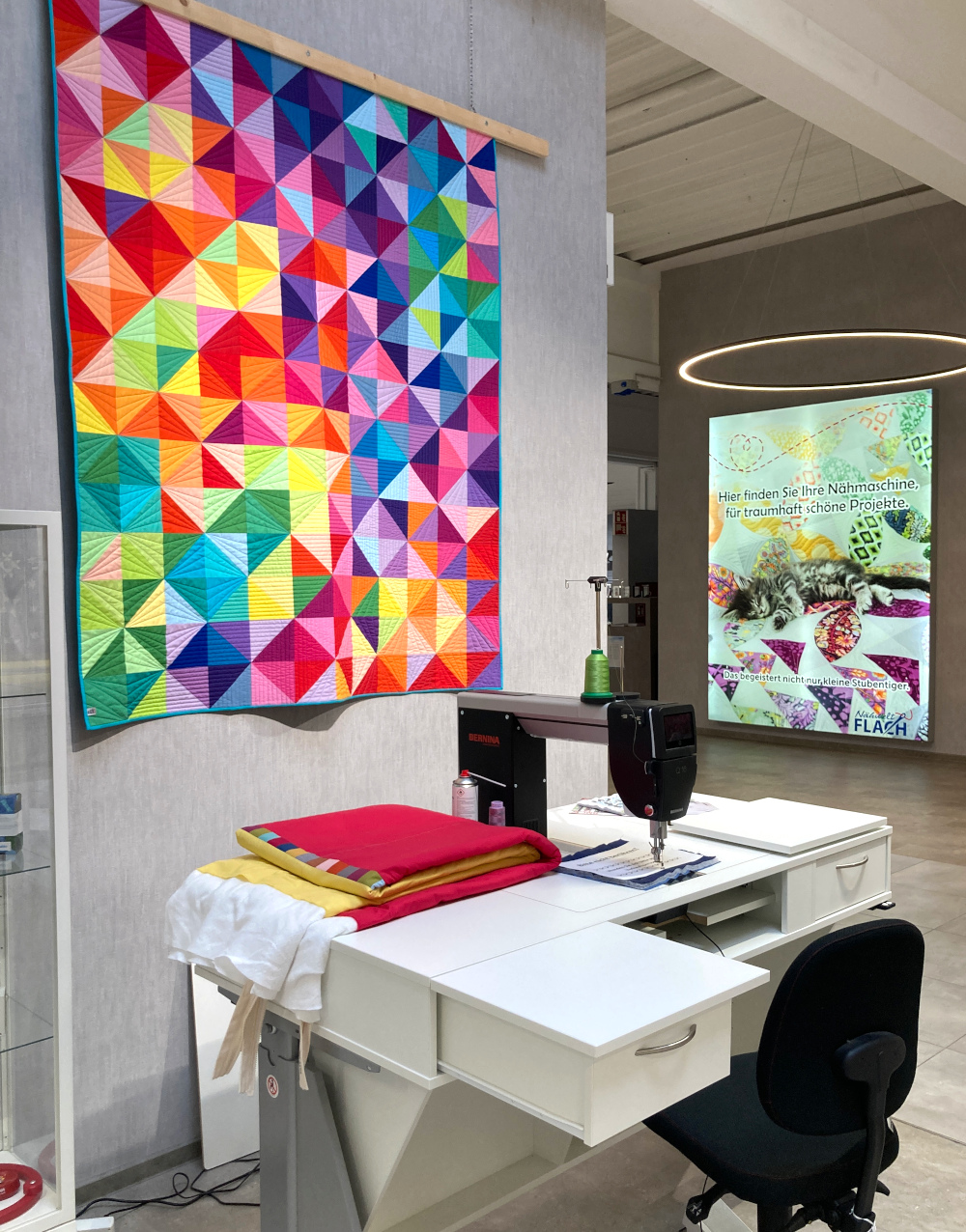 Im Ladengeschäft Nähwelt Flach: Vor einem bunten, geometrischen Wandquilt steht ein Tisch mit einer Nähmaschine. Davor steht ein schwarzer Drehstuhl.