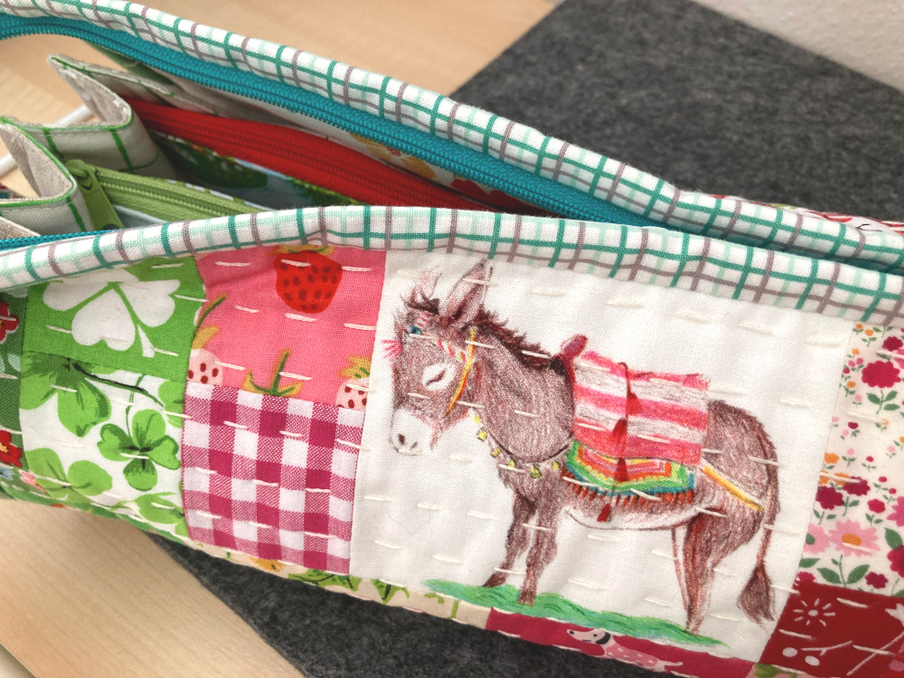 Halbgeöffnete Sew Together Bag aus grünen, pinken und weißen Stoffen, im Zentrum ein gezeichneter Esel mit gestreifter Decke auf dem Rücken
